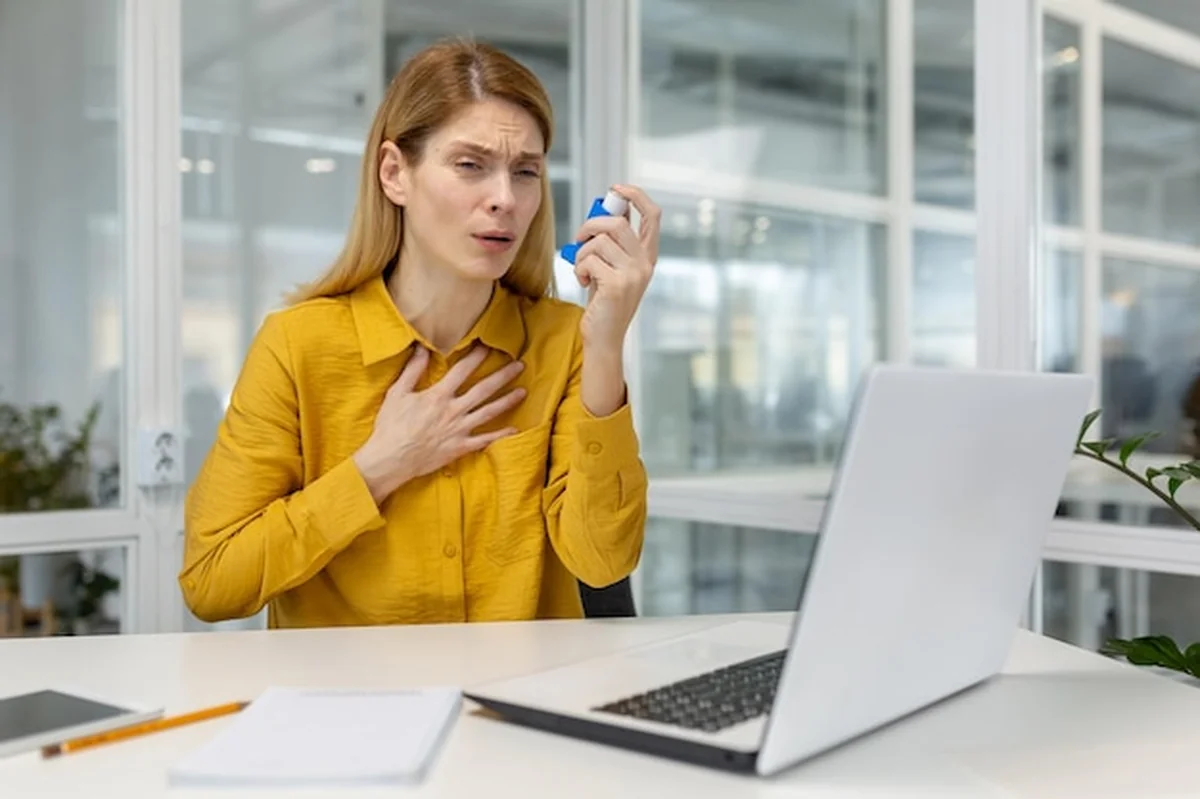 Kobieta z atakiem astmy używająca inhalatora przy laptopie w nowoczesnym biurze