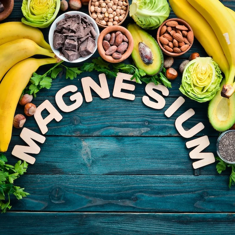 Pokarmy zawierające naturalny magnez: czekolada, banan, kakao, orzechy, awokado, brokuły, migdały na niebieskim tle